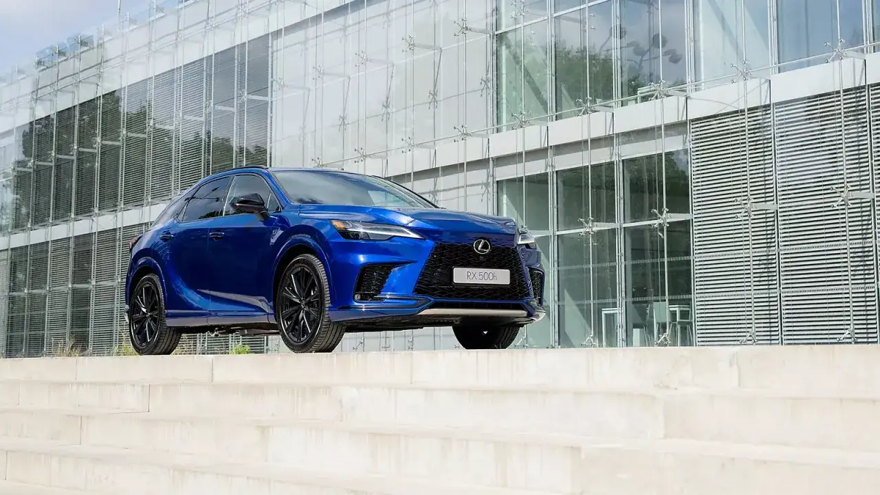 Błękitny Lexus RX500h zaparkowany na betonowych schodach przed nowoczesnym budynkiem z przeszkloną fasadą.