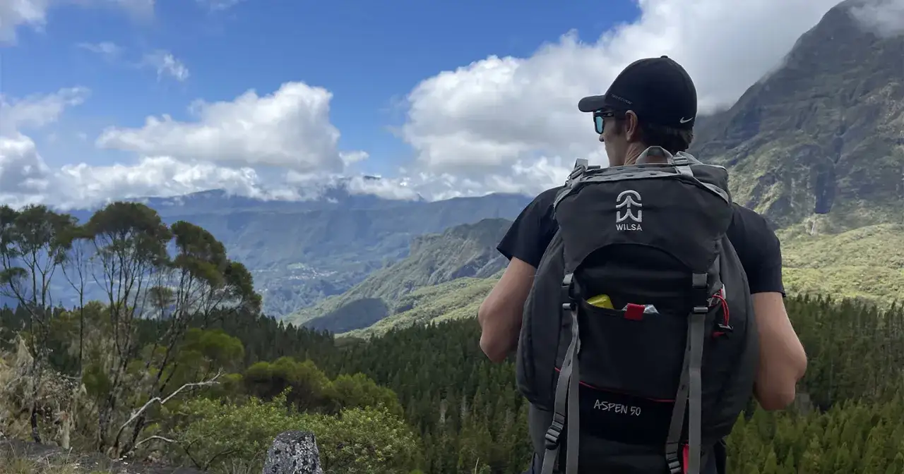 Un randonneur admire le paysage lors de son ascension d'une montagne. Le ciel est bleu avec des nuages blancs, et la vallée verdoyante s'étend à perte de vue.