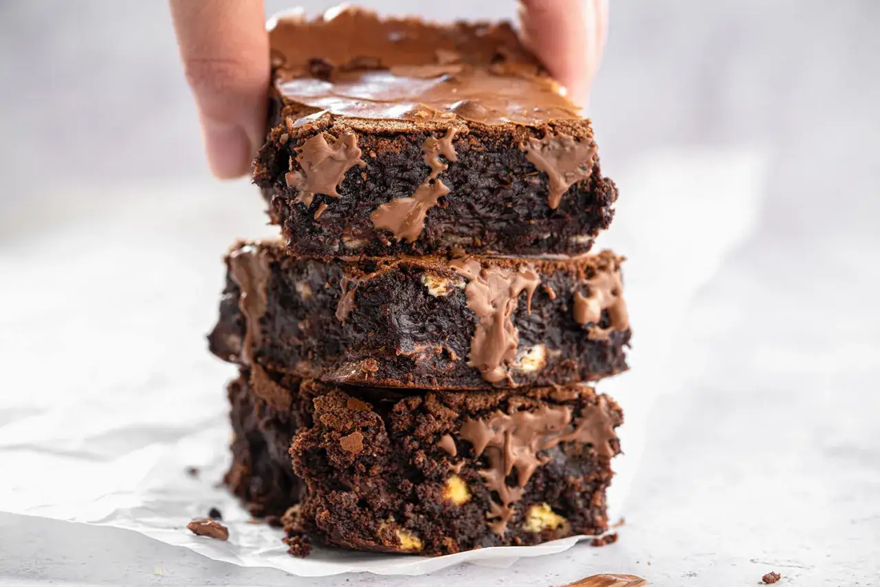 En hand lyfter en stapel saftiga, m&ouml;rka glutenfria brownies med sm&auml;lt choklad som rinner nerf&ouml;r sidorna.