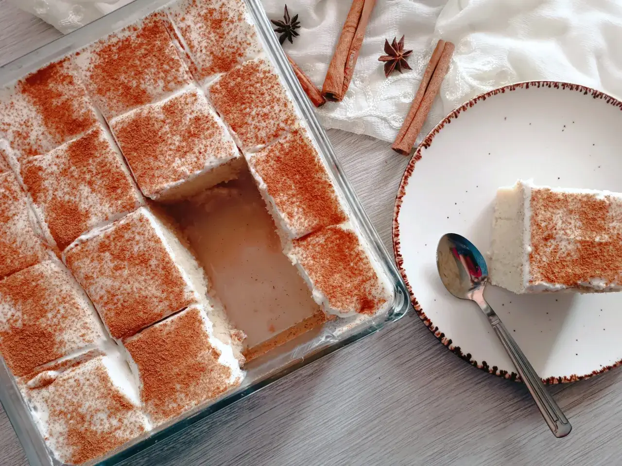 En saftig tres leches-kaka i en glasform, pudrad med kanel. En bit serveras på en tallrik med en sked.