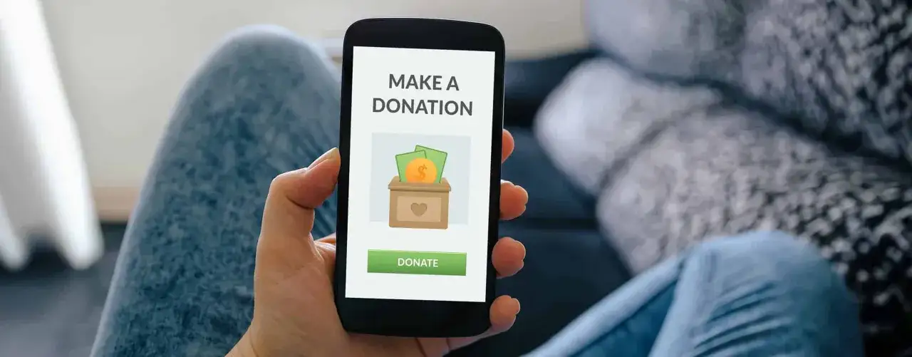 A person uses their phone for mobile donations to nonprofits. The screen shows a donation box with money and a heart icon.