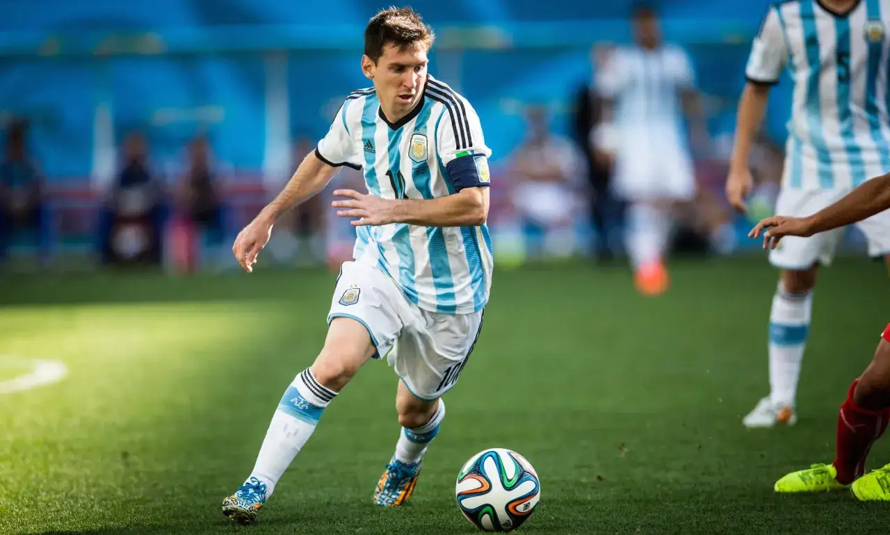 Leo Messi w białej koszulce z niebieskimi pasami, drybluje piłkę na zielonej murawie.