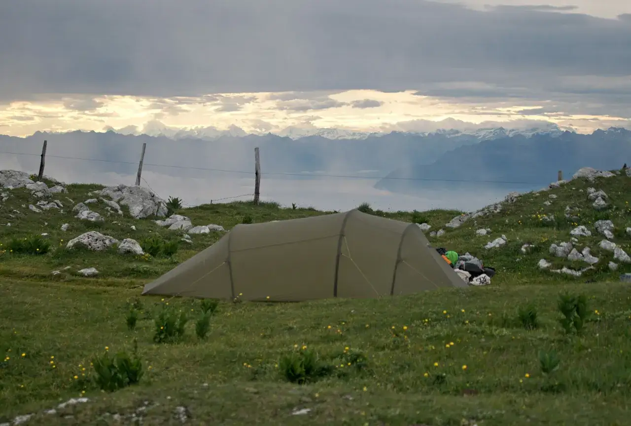 Ein Zelt steht auf einer gr&uuml;nen Wiese mit gelben Blumen. Im Hintergrund erheben sich majest&auml;tische Berge unter einem dramatischen Himmel. Camping in den Bergen.