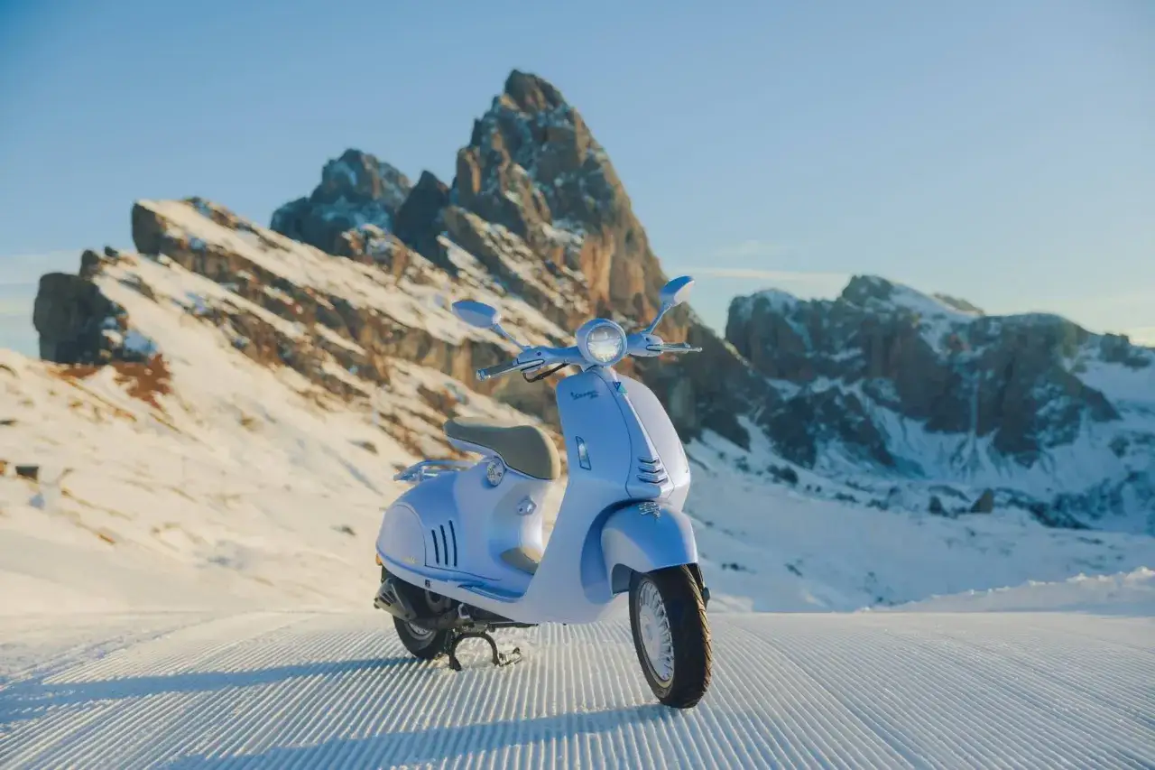 Una vespa azul marino descansa sobre nieve recién pisada, con imponentes montañas nevadas de fondo.