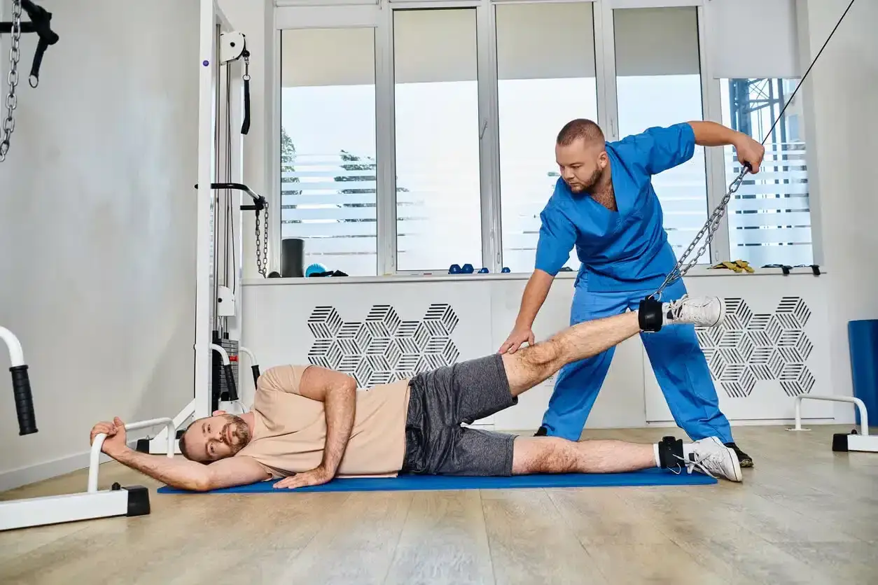 Mann macht Bein&uuml;bungen mit Gewichten, unterst&uuml;tzt von Therapeut. Wichtiger Schritt f&uuml;r Muskelaufbau nach Intensivstation.