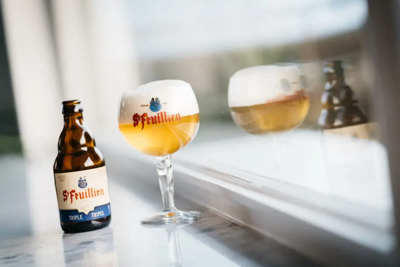 Une bouteille et un verre de bière de Bruxelles St-Feuillien Triple, avec un reflet dans la fenêtre.