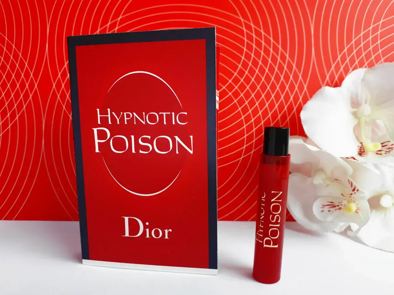 Czerwone opakowanie i flakonik perfum Christian Dior Hypnotic Poison na tle czerwonej, falistej tapety i białych kwiat&oacute;w.