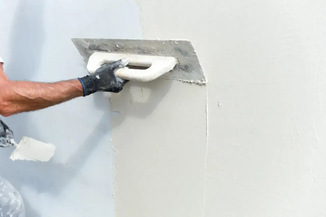 Mano enguantada aplicando estucado a pared con llana, creando una superficie lisa y uniforme.