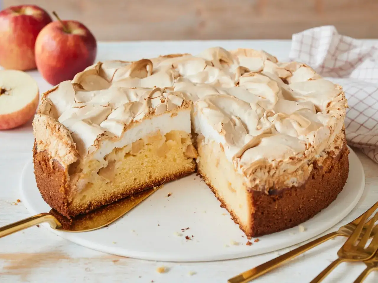 Ein saftiger Apfelkuchen mit Baiserhaube, der zum Anbei&szlig;en einl&auml;dt. Daneben liegen frische &Auml;pfel und goldenes Besteck.