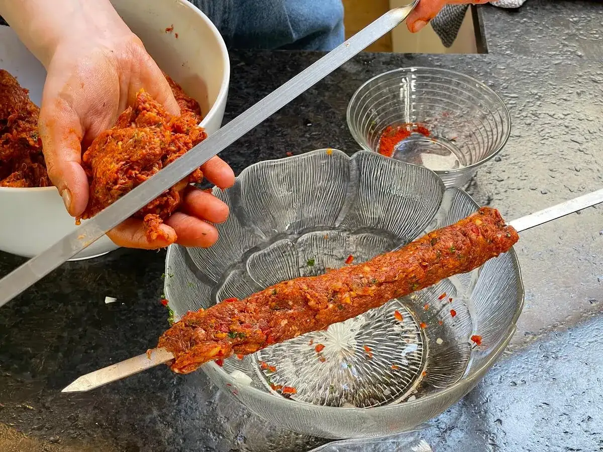 H&auml;nde formen Hackfleisch zu einem Adana Kebap auf einem Spie&szlig;. Gew&uuml;rze liegen bereit.