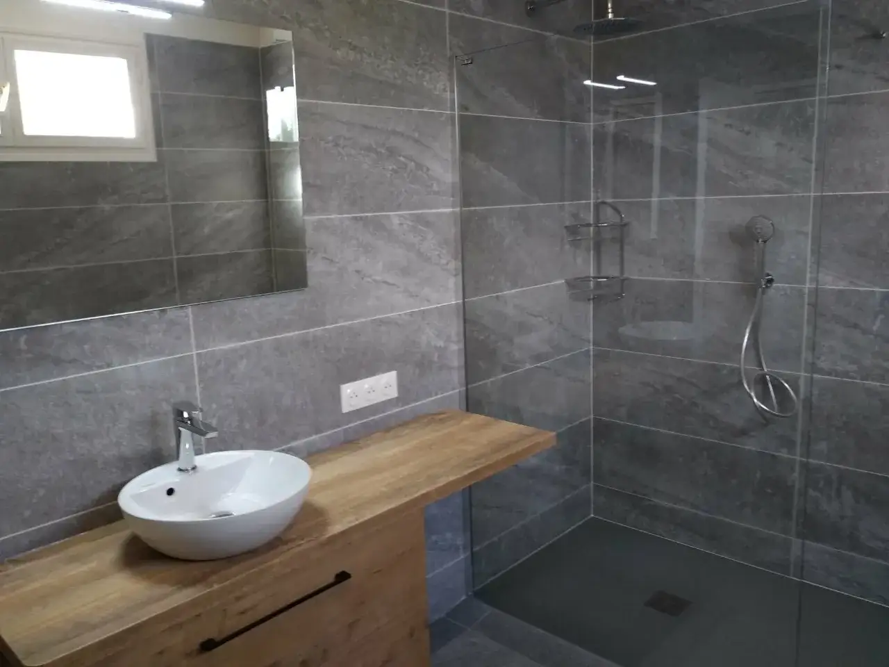 Salle de bain gris clair et gris fonc&eacute;. Douche moderne avec paroi en verre, lavabo sur meuble en bois, et miroir.