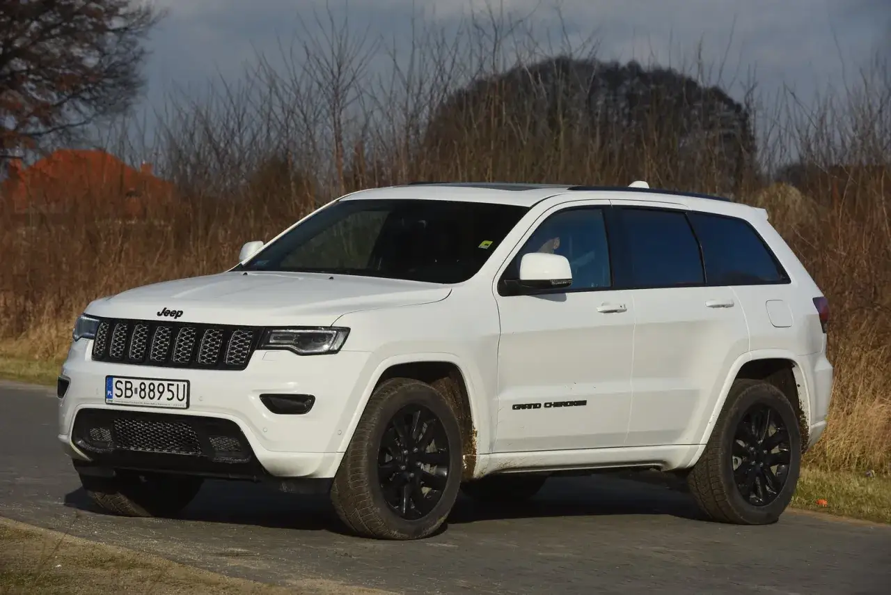 Biały Jeep Grand Cherokee 3.6 z czarnymi felgami, zaparkowany na tle zarośli i domu.
