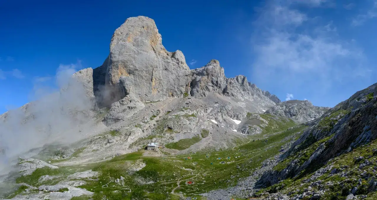 Majestuoso Naranjo de Bulnes, con refugio y tiendas de campaña a sus pies, bajo un cielo azul y nubes algodonosas.