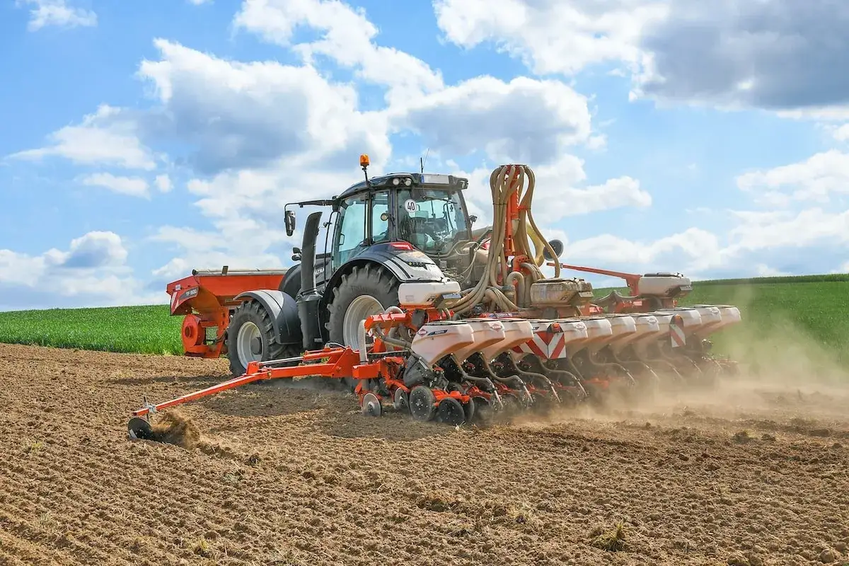 Trattore moderno impegnato nella coltivazione mais, semina in un campo arato sotto un cielo azzurro con nuvole bianche.
