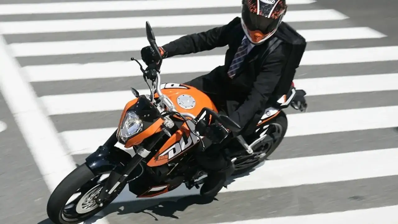 Elegancki mężczyzna w garniturze na pomarańczowym motocyklu KTM Duke 125, przejeżdża przez pasy. Dane techniczne tego modelu są imponujące.