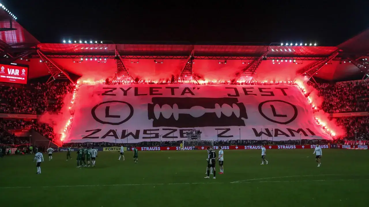 Trybuna stadionu rozświetlona racami, na środku ogromna biała płachta z napisem
