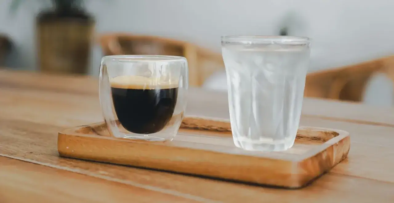 Ein Espresso und ein Glas Wasser auf einem Holztablett. Das Wasser dient dazu, den Gaumen zu reinigen, damit man den vollen Geschmack des Espressos genießen kann.