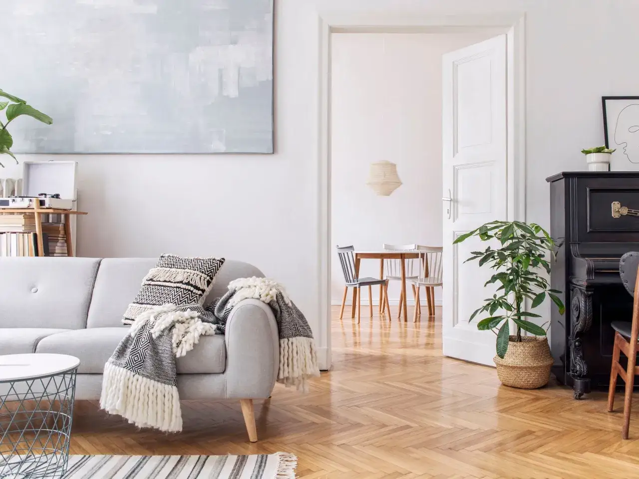 Sal&oacute;n acogedor con sof&aacute; gris, manta de punto y coj&iacute;n decorativo. Al fondo, un comedor con mesa de madera y sillas. Iluminaci&oacute;n c&aacute;lida de 4000 K.