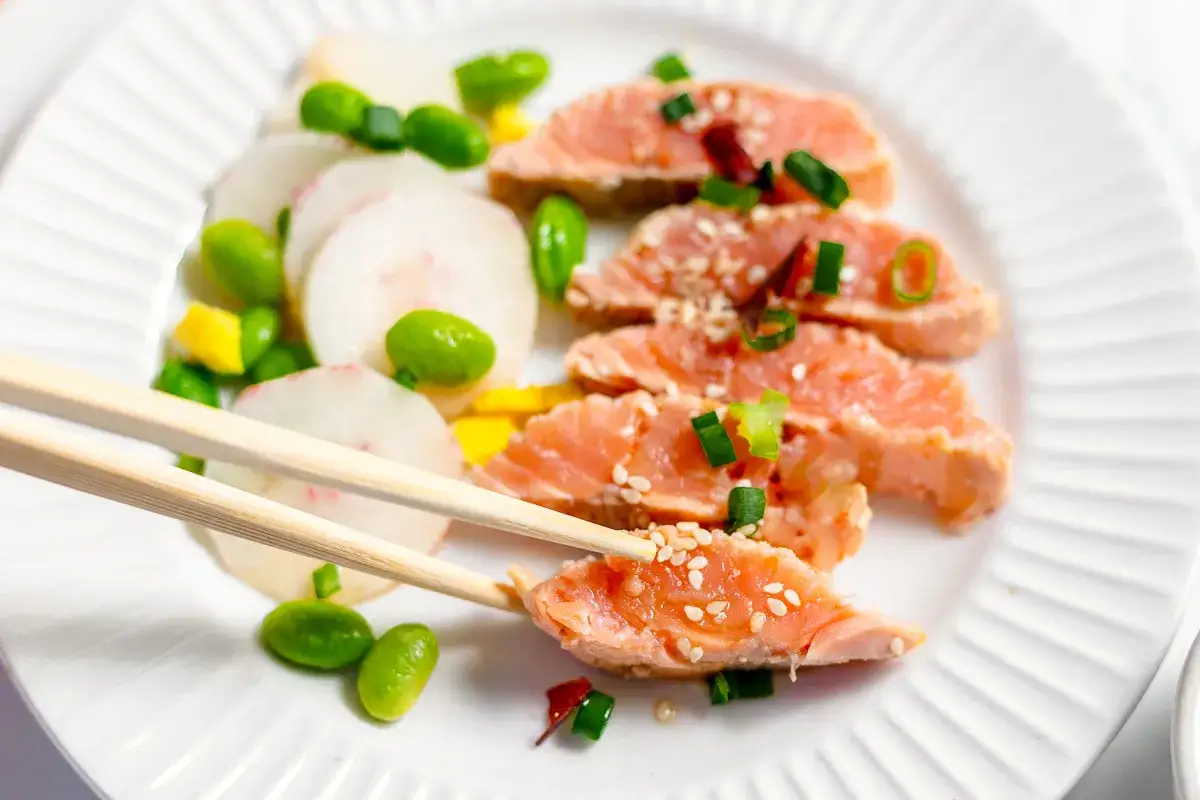 Tataki de salmón con rodajas de rábano, edamame y cebolleta. Palillos recogen un trozo.