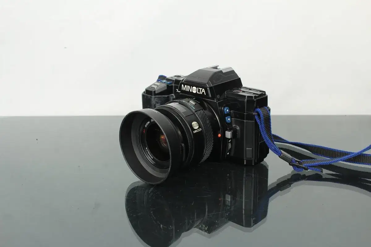 Svart Minolta 7000 systemkamera med objektiv och bl&aring; axelrem, placerad p&aring; en reflekterande yta.