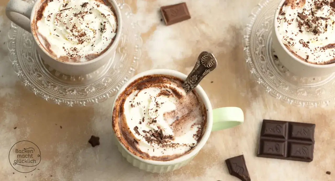 Drei Tassen mit cremigem, heißem Kakao, garniert mit Schlagsahne und Schokoladenraspeln. Ein Löffel ruht in einer Tasse, während Schokoladenstücke daneben liegen.