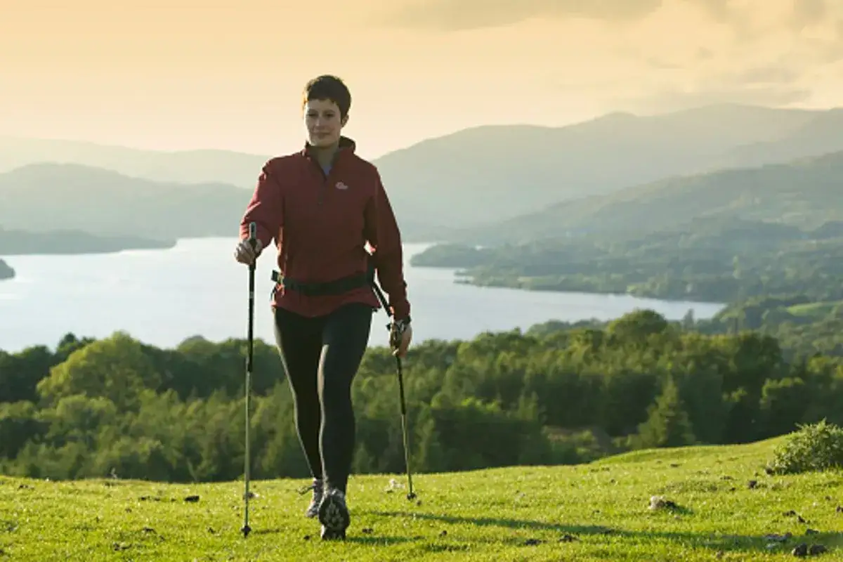 Kobieta z kijkami do nordic walking na tle gór i jeziora. Widać wyraźne efekty aktywności fizycznej.