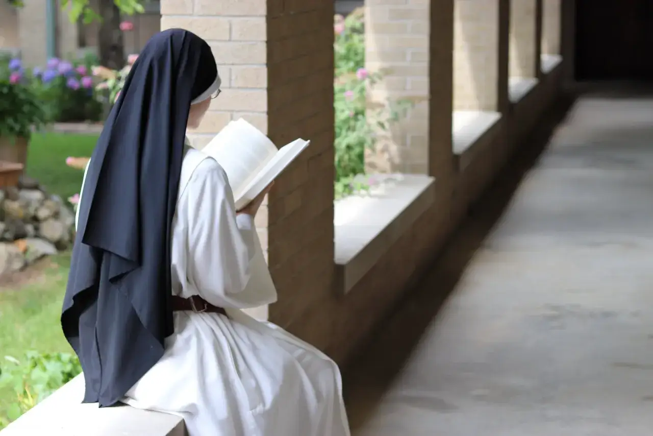 A nun in a white habit and black veil reads a book, embodying monk characteristics of contemplation and study.