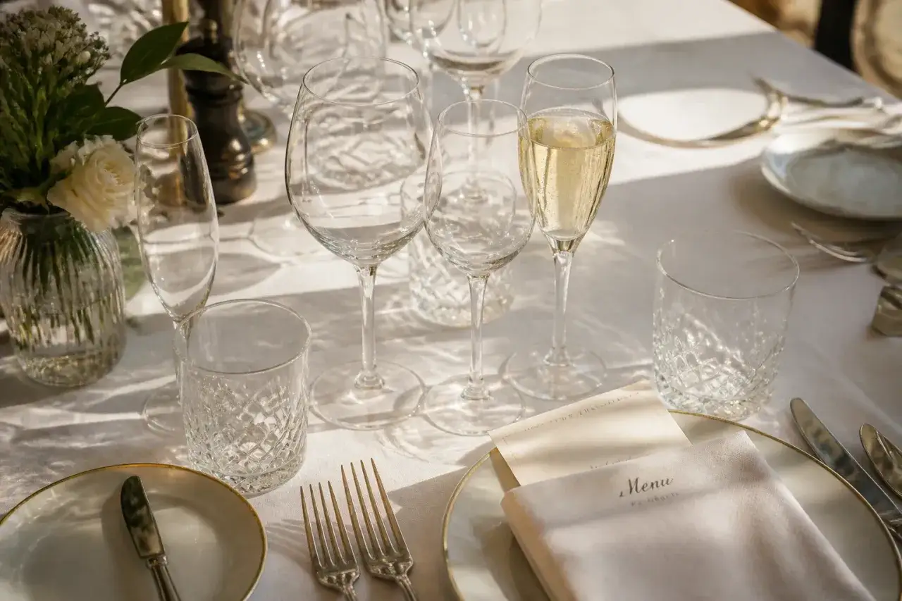 Une table dressée pour un repas, avec des verres de différentes formes, un menu et des couverts. Une histoire du verre à travers ces objets élégants.