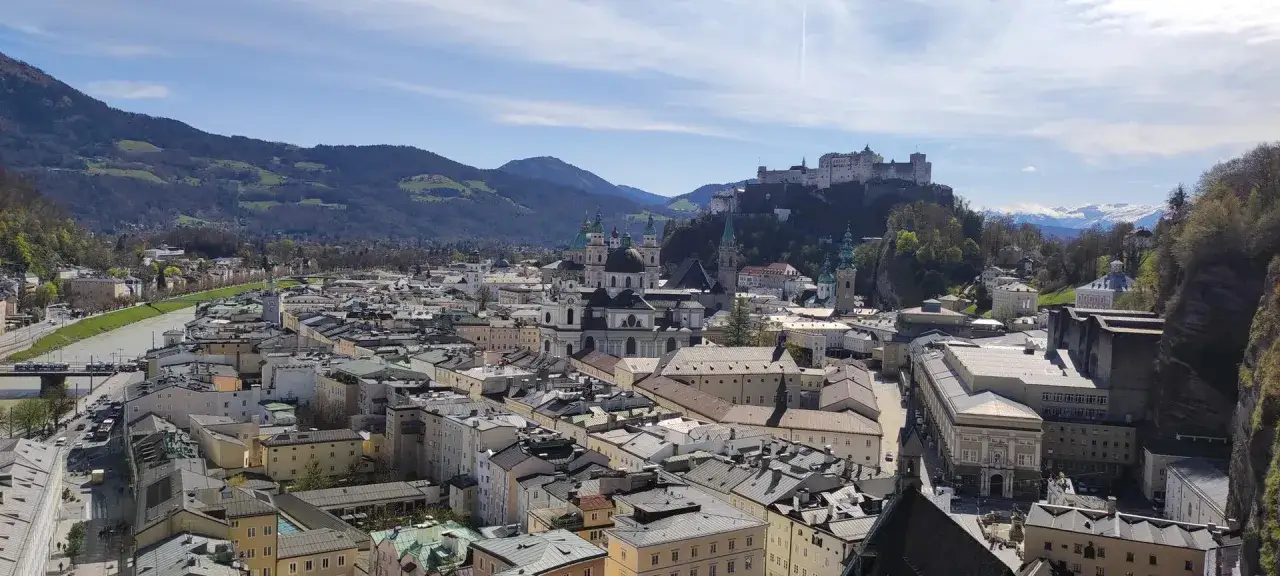 Blick über Salzburg mit Festung Hohensalzburg und dem Kapuzinerkloster. Berge und blauer Himmel im Hintergrund.