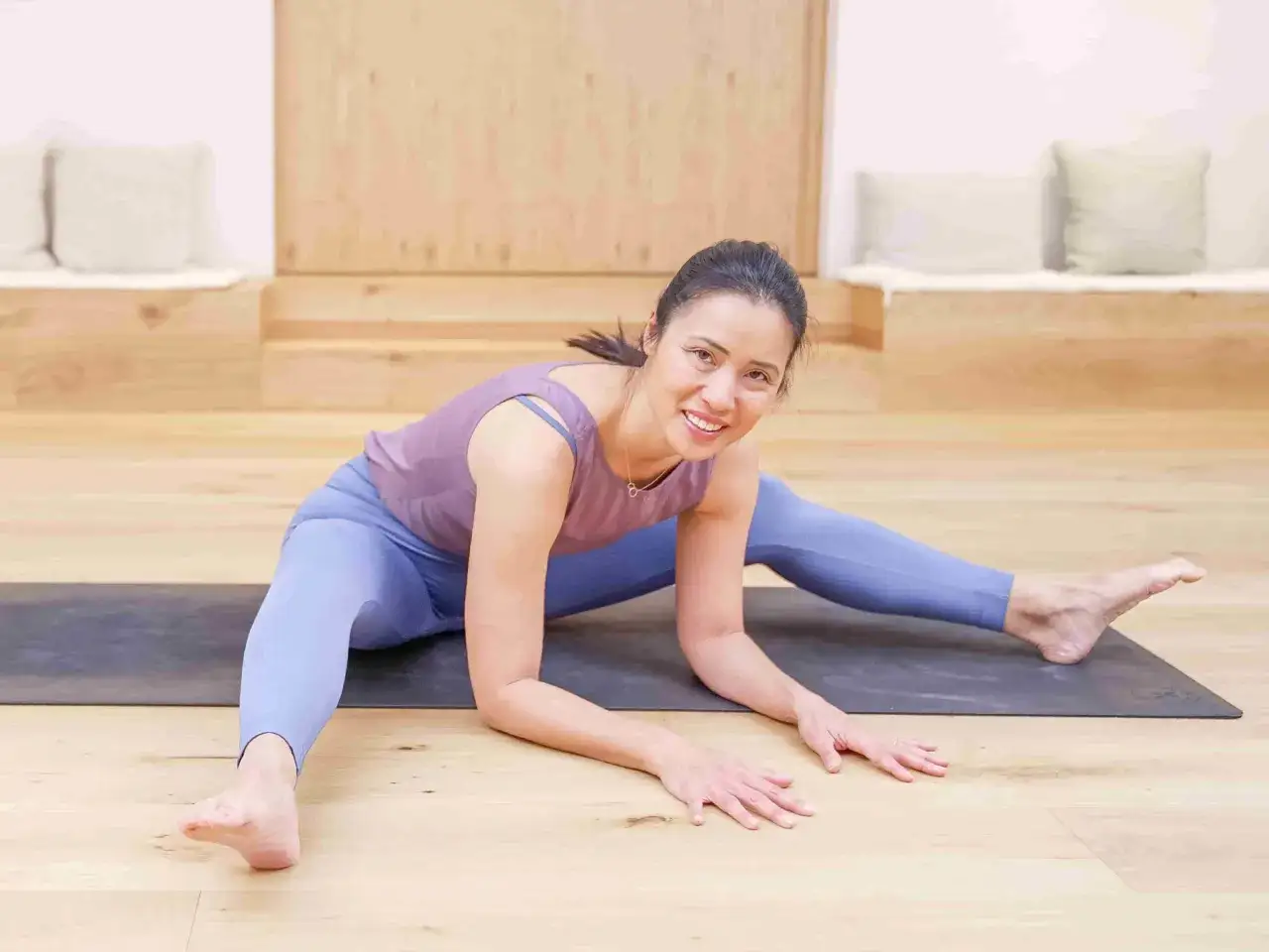 Mujer sonríe en postura de yoga, con piernas abiertas y brazos apoyados en el suelo. Su práctica de karma yoga la llena de paz.