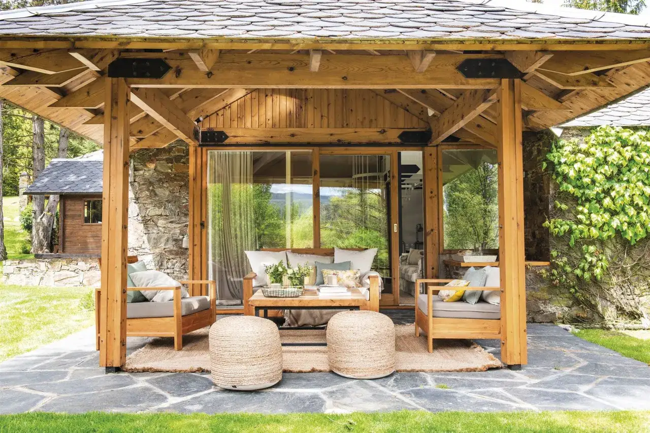 Porches de casas sencillas con mobiliario de madera y cojines, alfombra de yute y pufs. Ventanales que dan a un paisaje verde.
