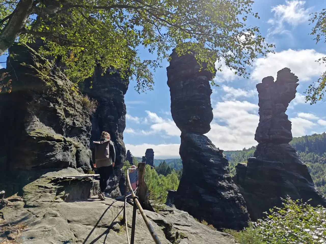 Wanderer mit Rucksack auf einem Felsenpfad im Bielatal, umgeben von bizarren Sandsteinfelsen und gr&uuml;nen B&auml;umen.