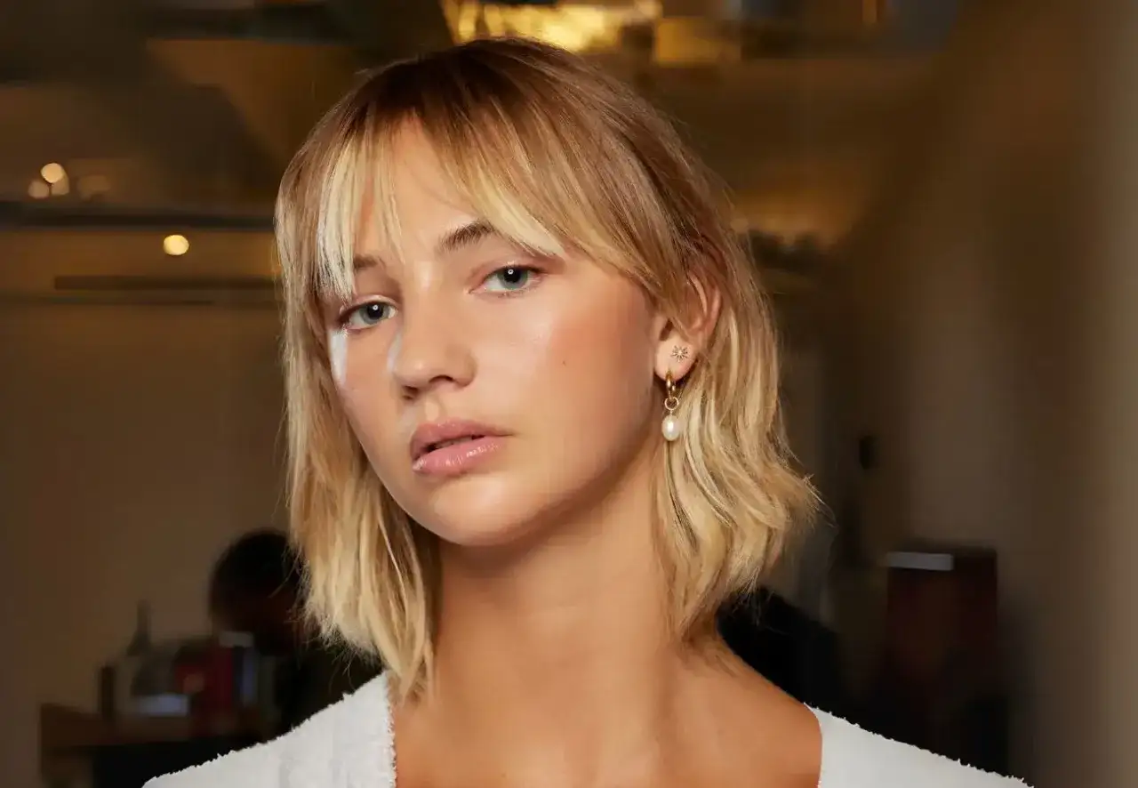 Mujer joven con un corte long bob rubio y flequillo. Luce un pendiente de perla y estrella.