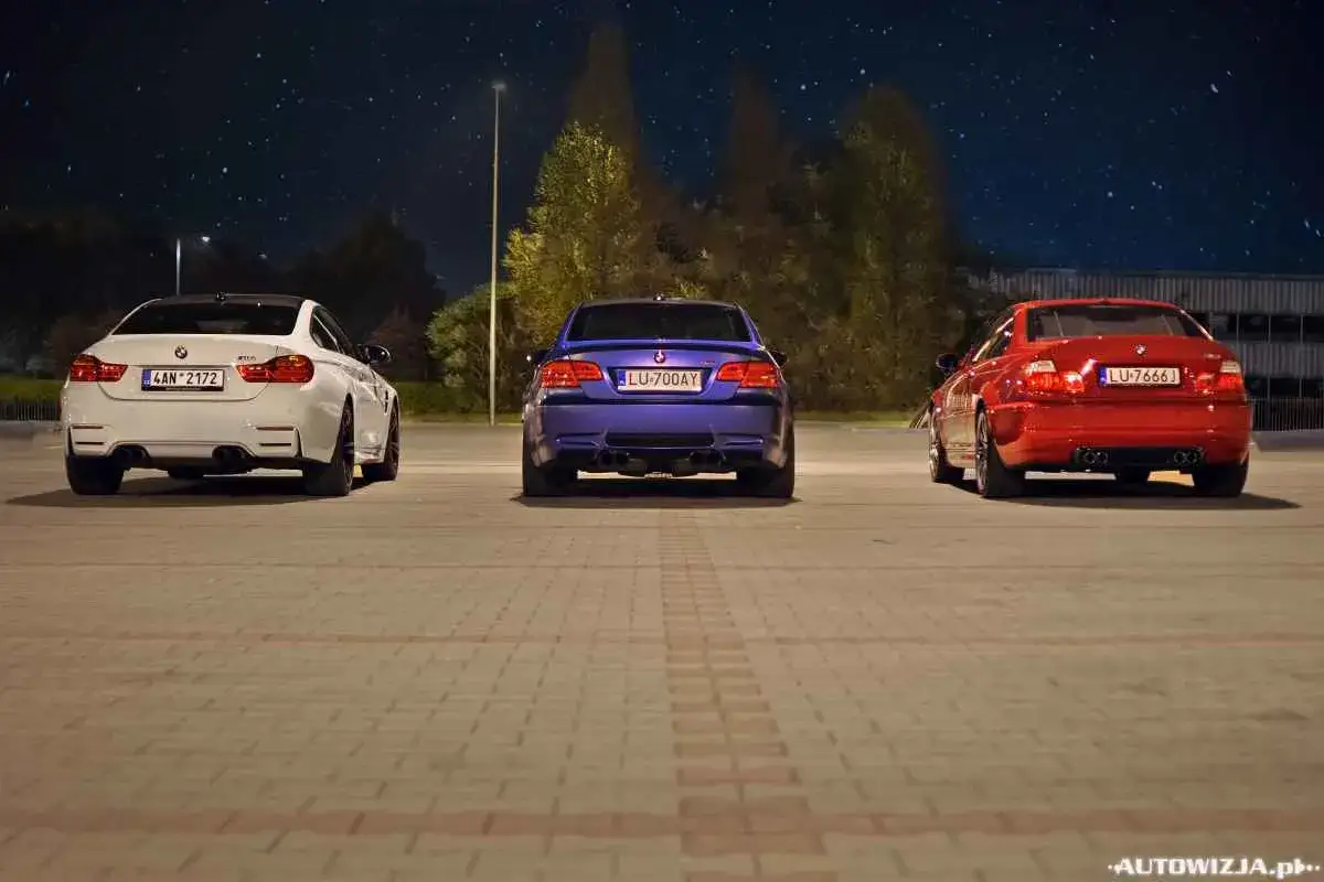Trzy BMW: białe, niebieskie i czerwone, stoją nocą na parkingu. Niebo pełne gwiazd nad nimi.