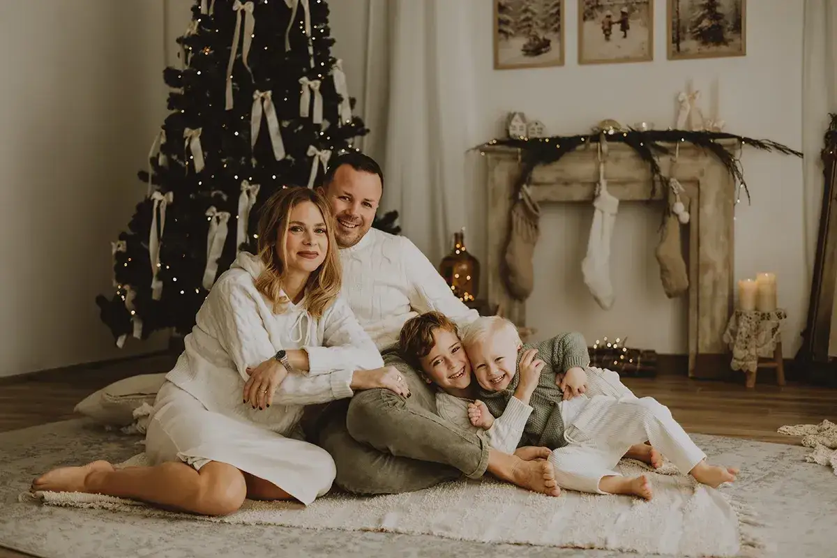 Una dolce fotografia natalizia: una famiglia sorride felice davanti all'albero addobbato e al camino con le calze appese.