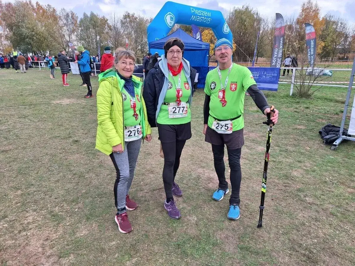 Trzy osoby w strojach sportowych, gotowe do startu w zawodach nordic walking. Uśmiechnięci uczestnicy z numerami startowymi.