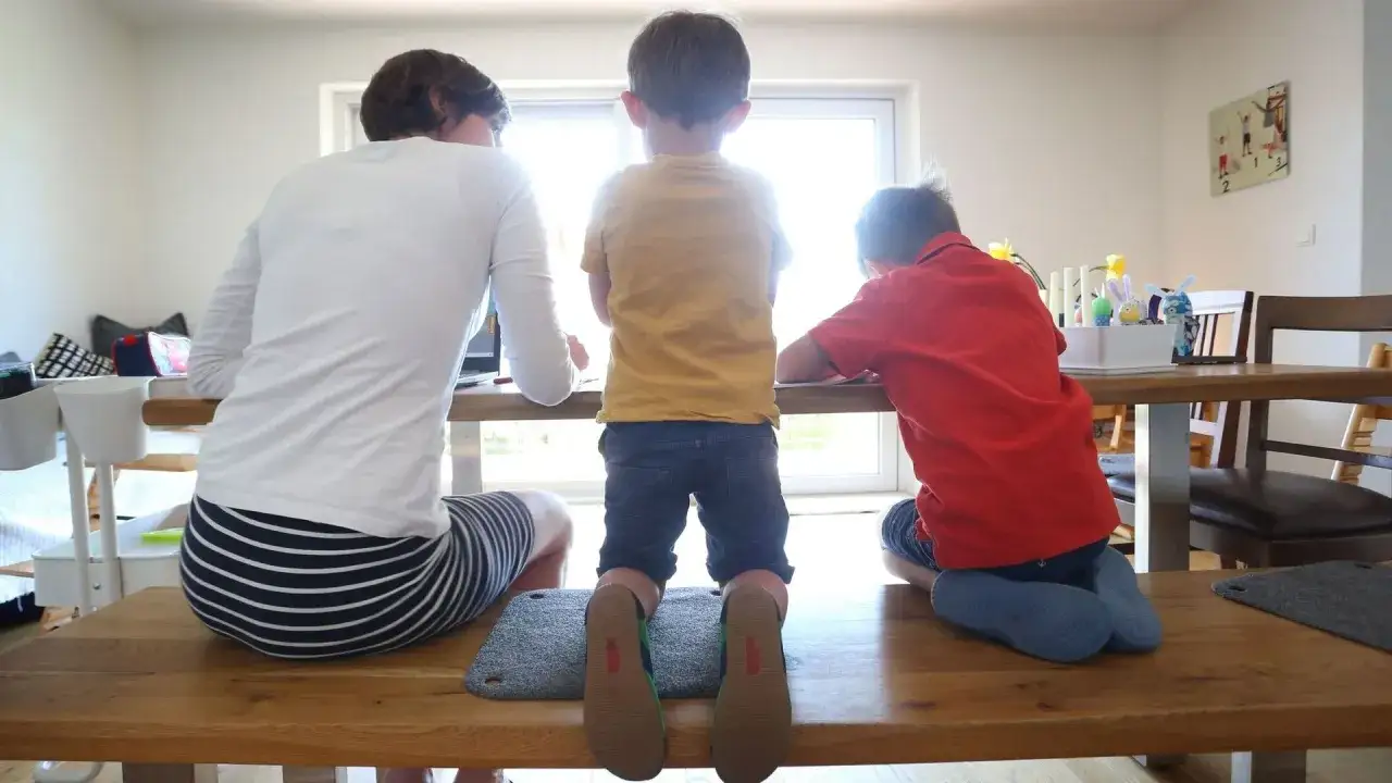 Eine Frau und zwei Kinder sitzen an einem Tisch. Die Frau ist mit ihrer Arbeit beschäftigt, während die Kinder malen. Die Szene könnte eine toxische Tochter darstellen, die ihre Mutter ignoriert.