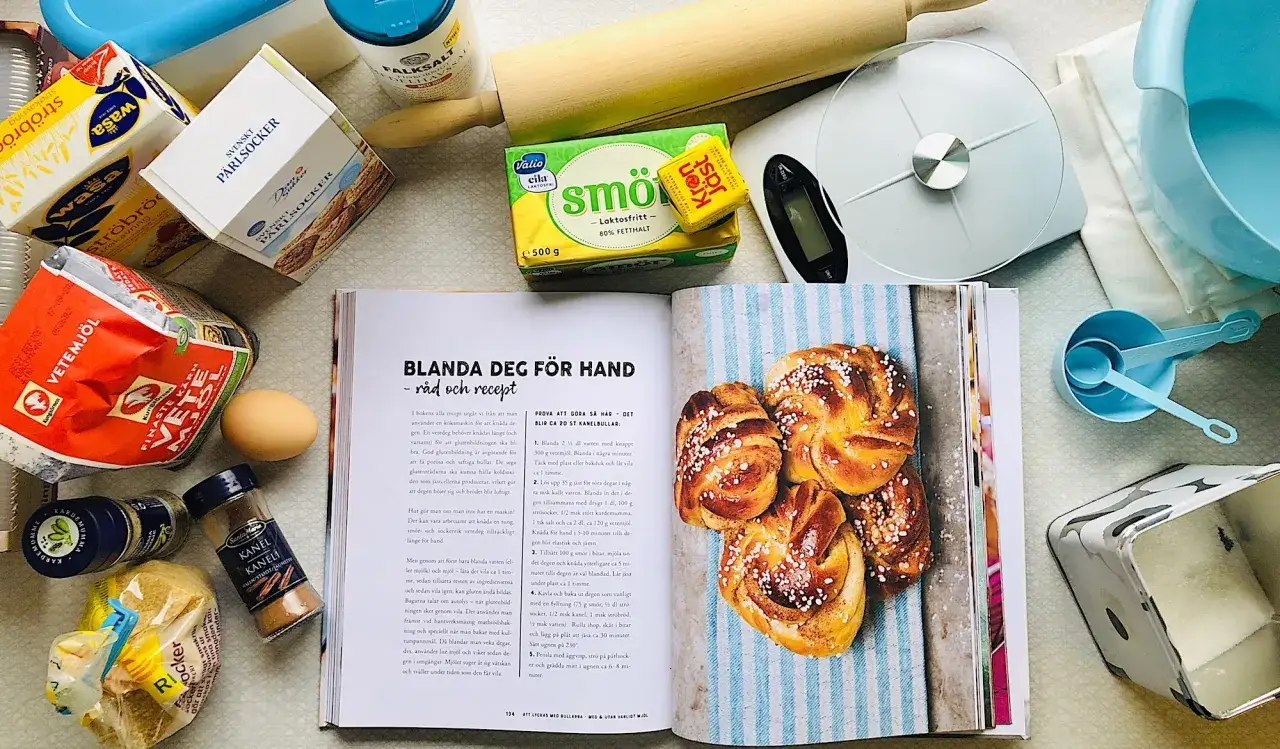 Här ligger ingredienser för att baka med söt jäst, som vetemjöl, socker, smör och jäst. En öppen bok visar recept och kanelbullar.