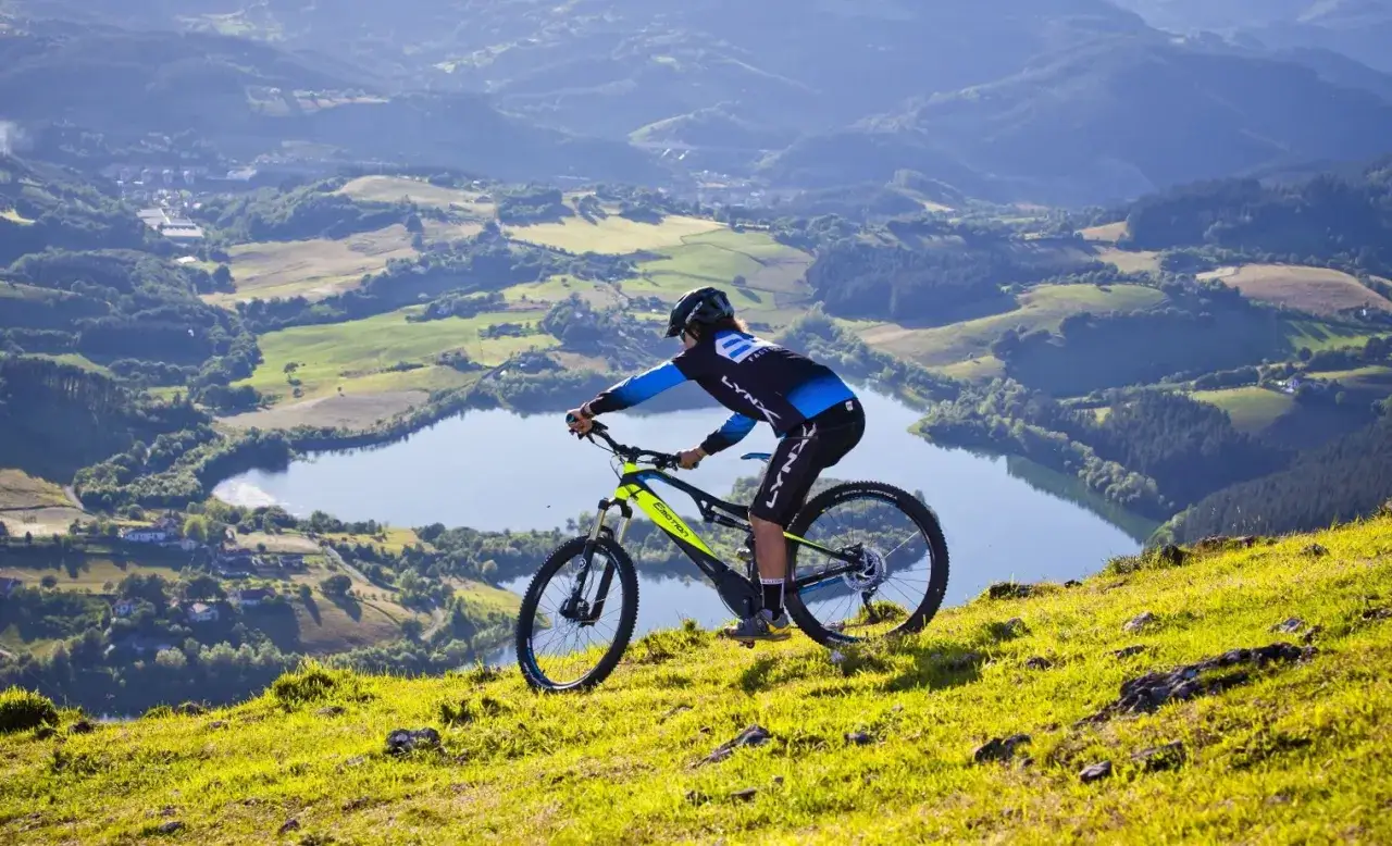 Ciclista desciende por una ladera verde, disfrutando de las vistas de un lago y montañas. ¡Perfecto para rutas bici Valencia!