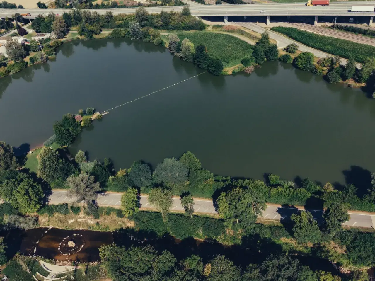Luftaufnahme vom Badesee Waldhausen mit einer schwimmenden Boje-Linie, einer kleinen Insel mit H&uuml;tte und einer Stra&szlig;e am Ufer.