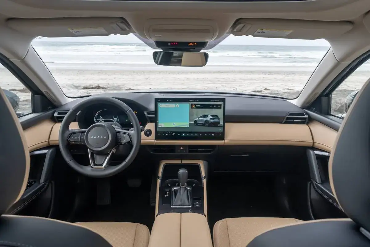 Wnętrze CX-5 Mazdy z widokiem na plażę. Kierownica, ekran multimedialny z nawigacją i jasna tapicerka tworzą komfortową przestrzeń.