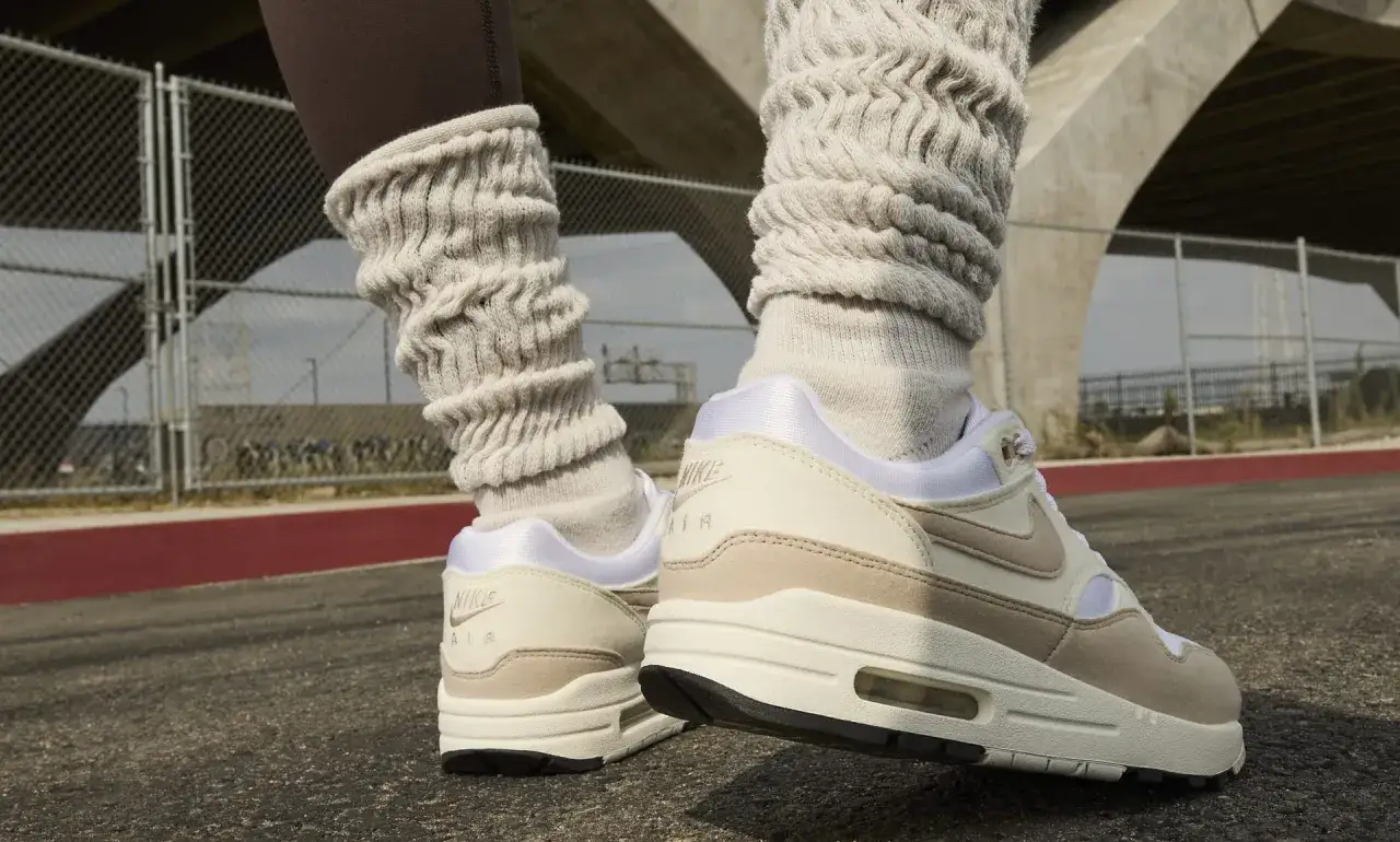 Nogi w beżowych skarpetach i podkolanówkach, na nogach buty Nike Air Max Light w kremowo-beżowej kolorystyce, na tle betonowego mostu.