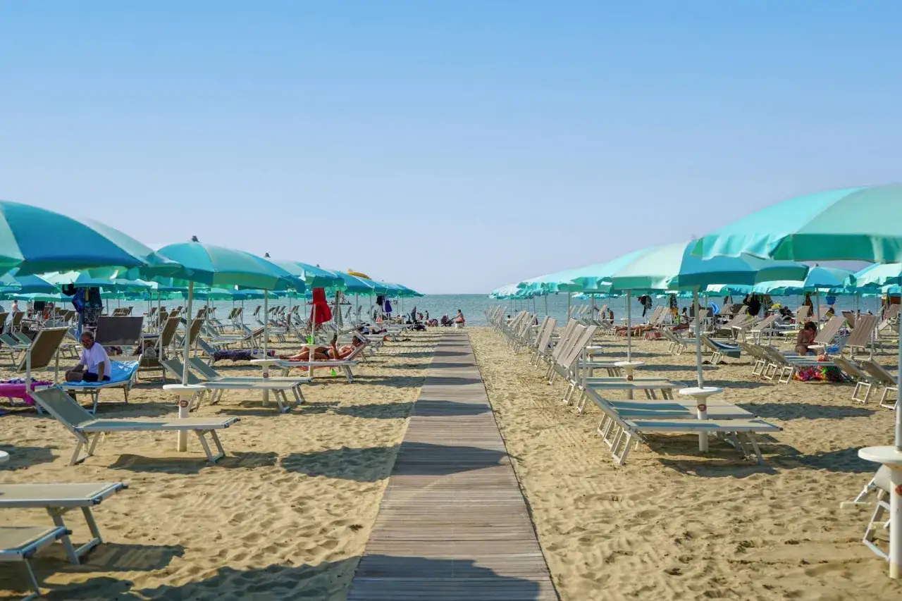 Una passerella di legno conduce alla spiaggia di San Giuliano Mare, fiancheggiata da file di lettini e ombrelloni azzurri sotto un cielo limpido.