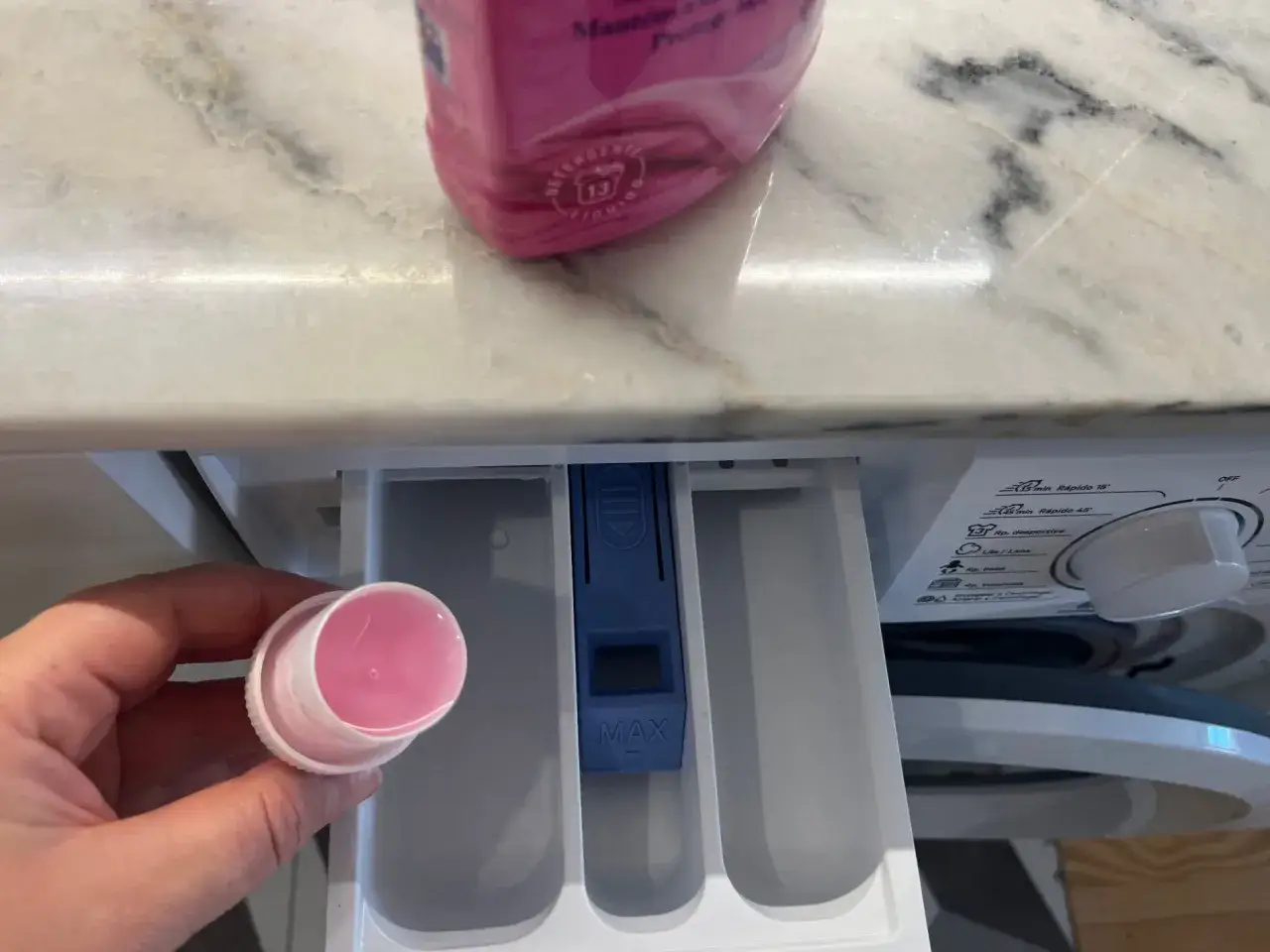 Hand h&auml;lt Messbecher mit rosa Fl&uuml;ssigkeit, bereit, Waschpulver direkt in die Trommel zu geben. Waschmittelschublade ist ge&ouml;ffnet.