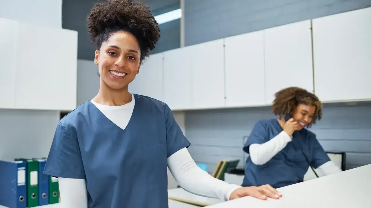 Secr&eacute;taire m&eacute;dicale souriante, pr&ecirc;te pour sa formation en ligne. Une coll&egrave;gue au t&eacute;l&eacute;phone en arri&egrave;re-plan.
