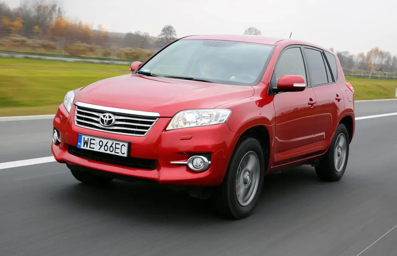 Czerwony SUV do 30 tys. jedzie po drodze, za nim zielone pola i drzewa.