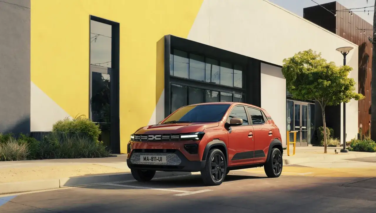 Dacia Evader arancione parcheggiata davanti a un edificio moderno con pareti gialle e bianche.