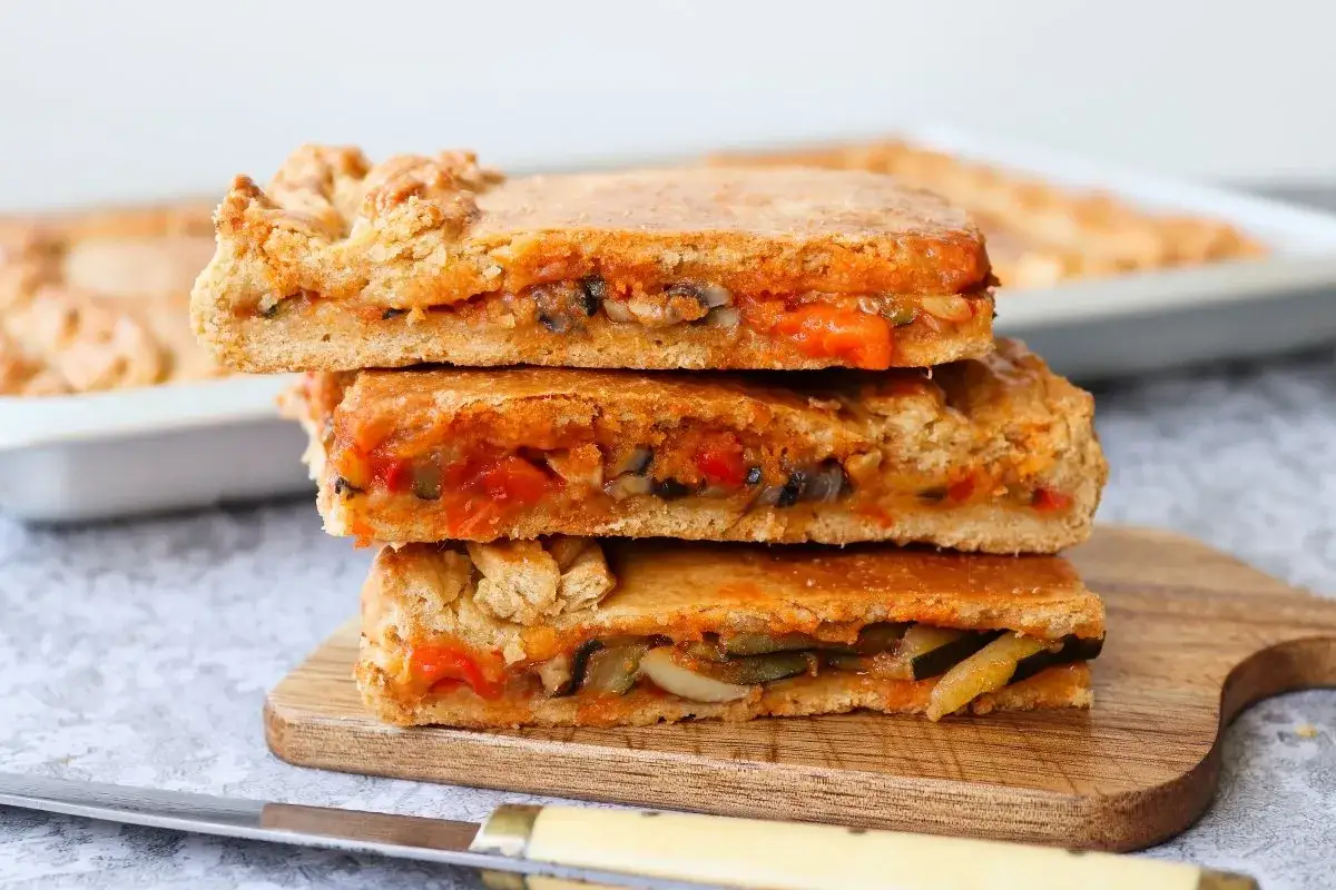 Tres porciones apiladas de empanada de verduras, rellenas de pimientos, calabacín y tomate.