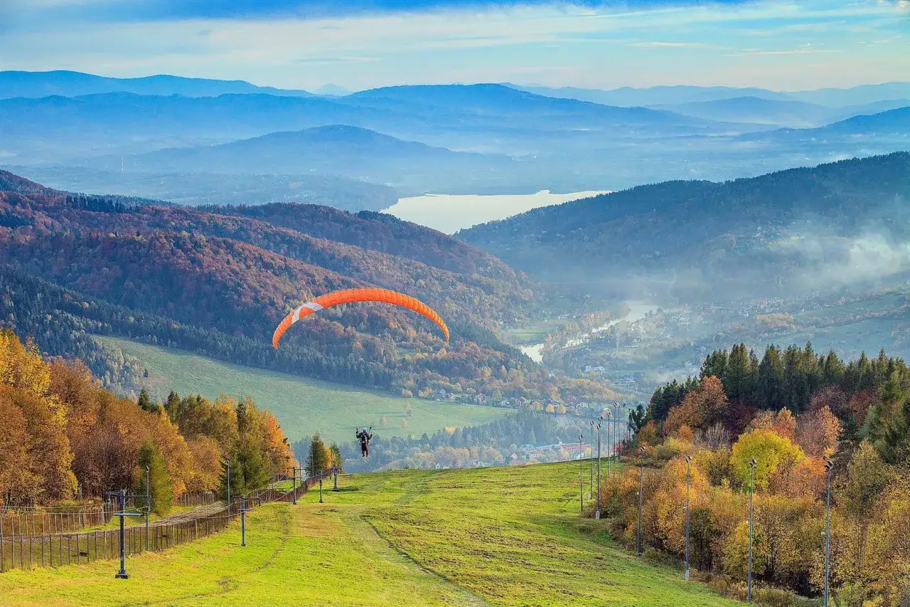Pomarańczowy paralotniarz szybuje nad jesienną trasą góry Żar, podziwiając malowniczą dolinę i jezioro.
