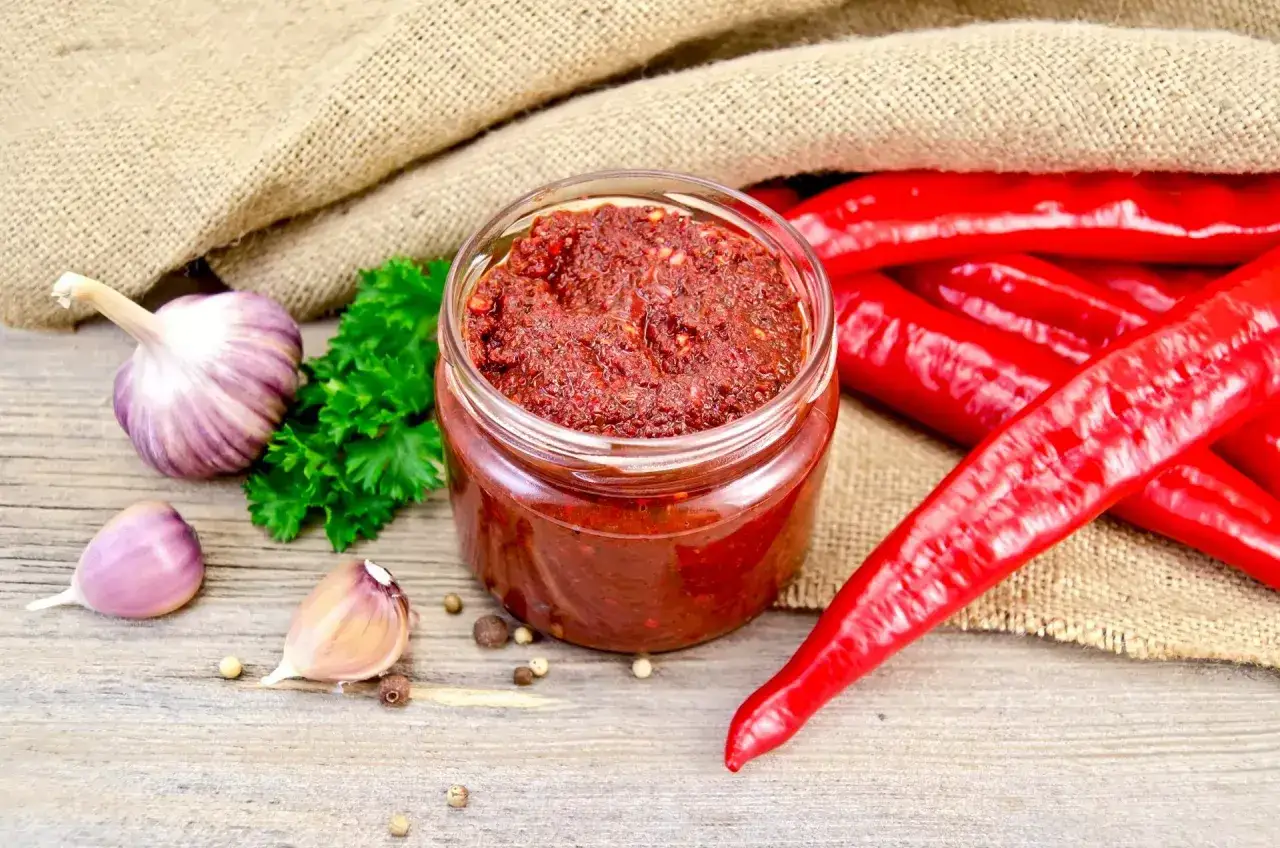 Słoik pełen pikantnego sosu adżika, obok świeży czosnek, natka pietruszki i czerwone papryczki chili.